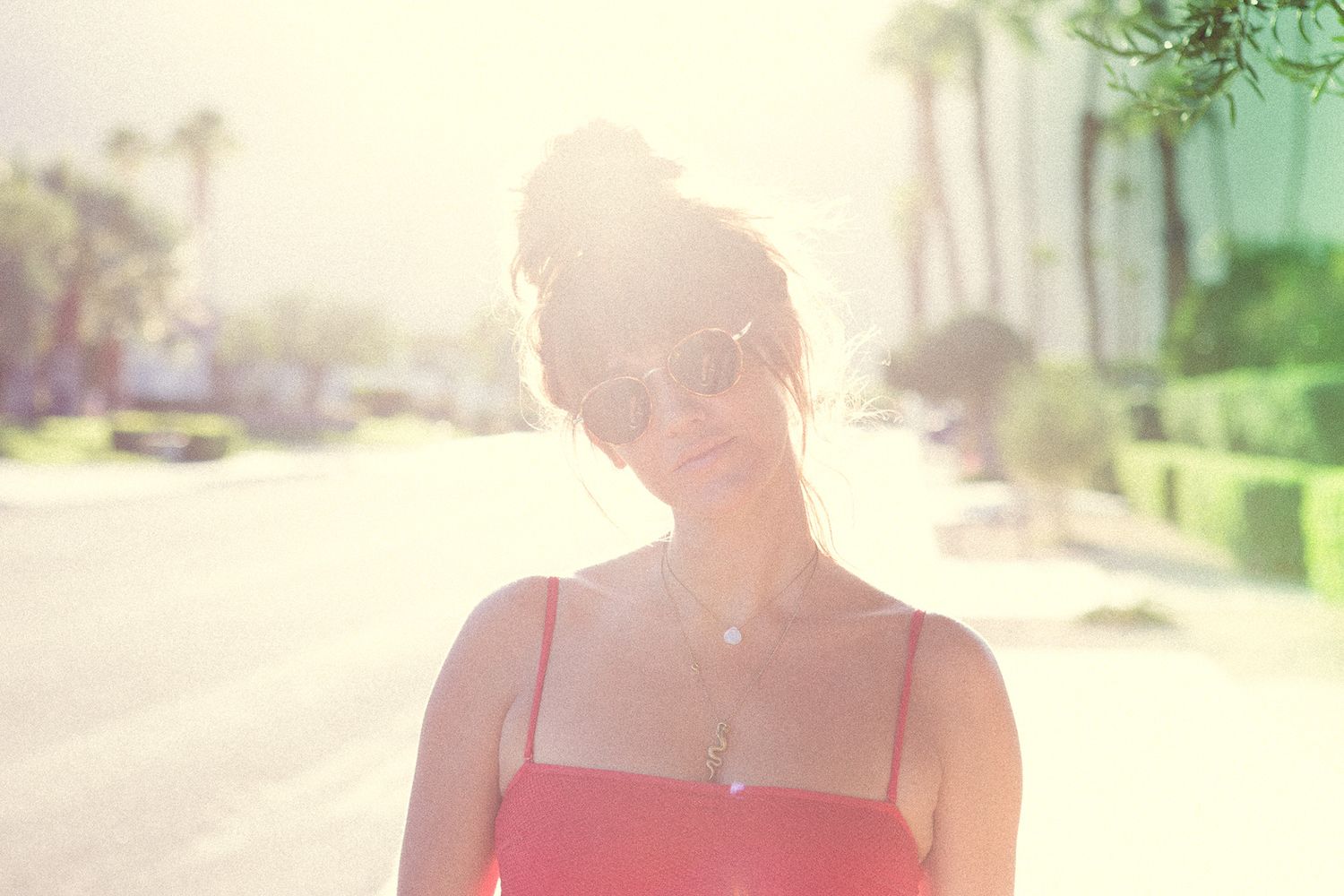 Young woman in a red dress and sunglasses standing outdoors in bright sunlight near palm trees.