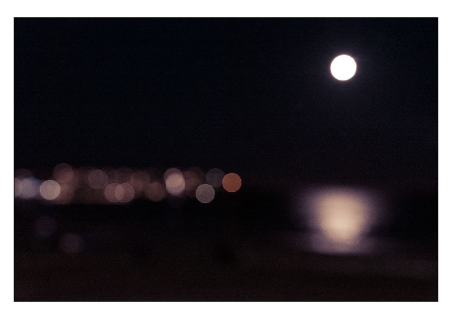 Blurry night photo of a full moon reflecting over water with bokeh city lights.