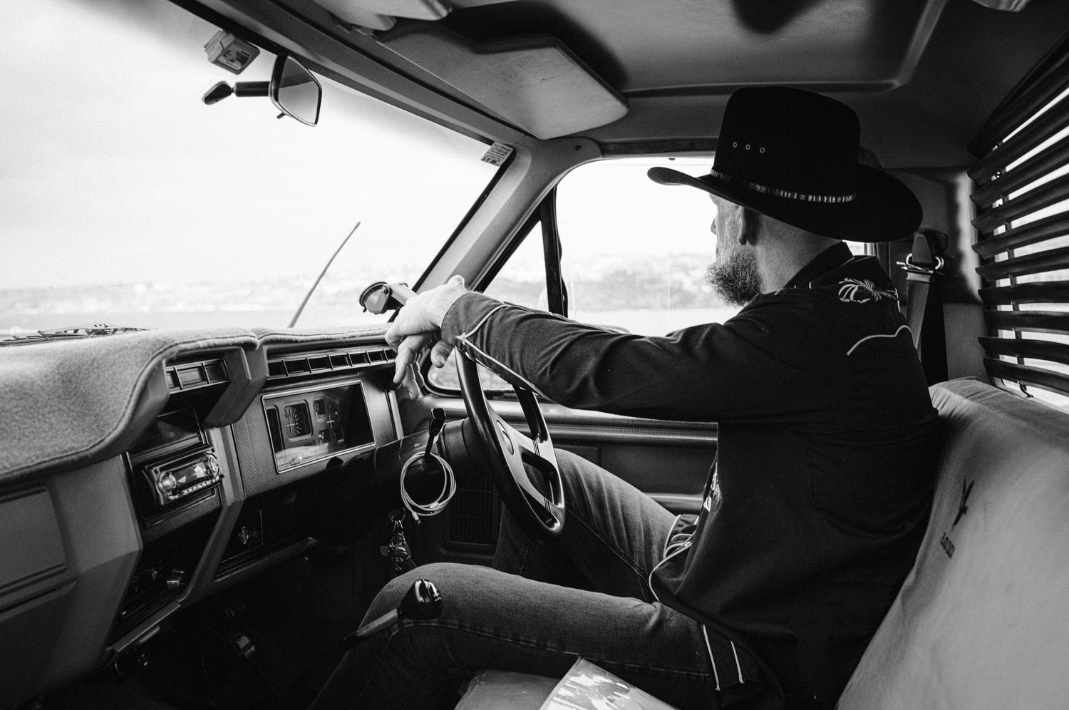 Man in black cowboy hat driving vintage truck on open road, black and white.