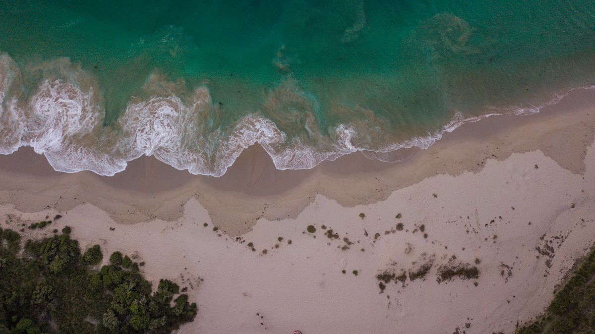 Aerial view of turquoise ocean waves gently washing onto a pristine sandy tropical beach with lush green trees.