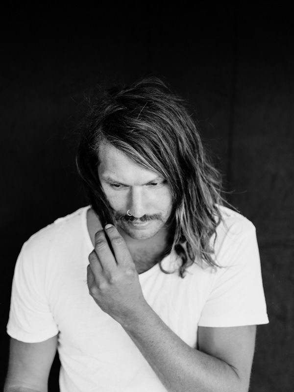 Black and white portrait of long-haired man with nose ring in white t-shirt looking down.