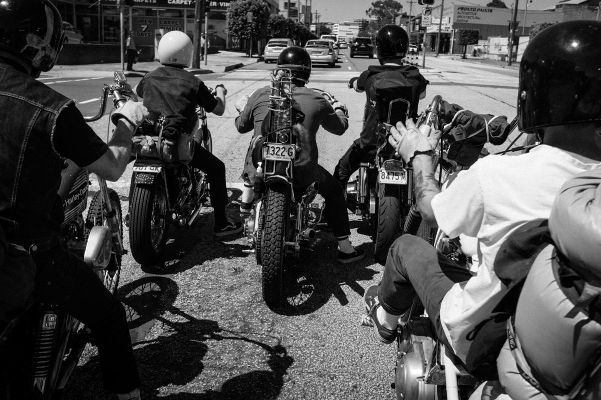 Black and white photo of a group of motorcyclists gathered on a sunny street.