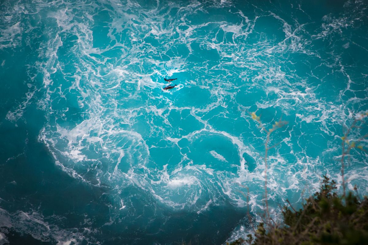 Aerial view of swirling turquoise ocean waves crashing against a rocky coastline with white sea foam.
