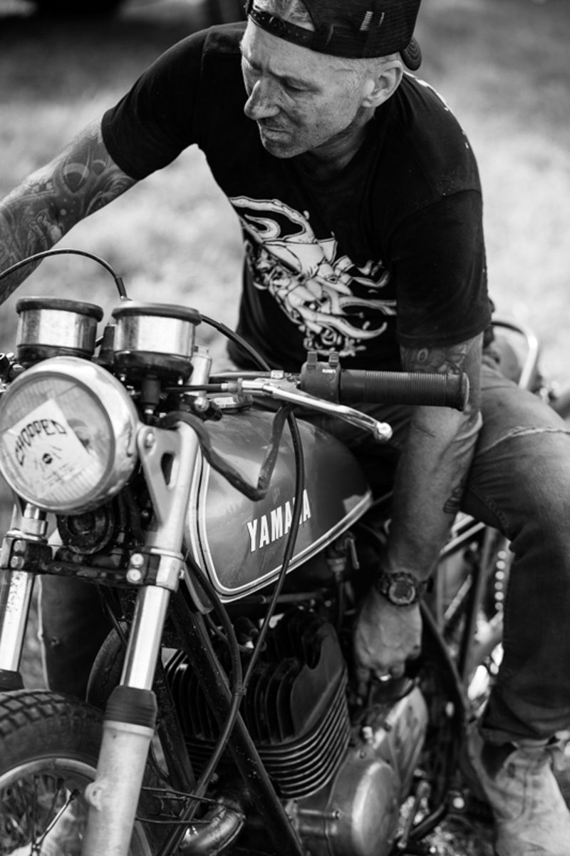 Black and white photo of a tattooed man sitting on a vintage Yamaha motorcycle.