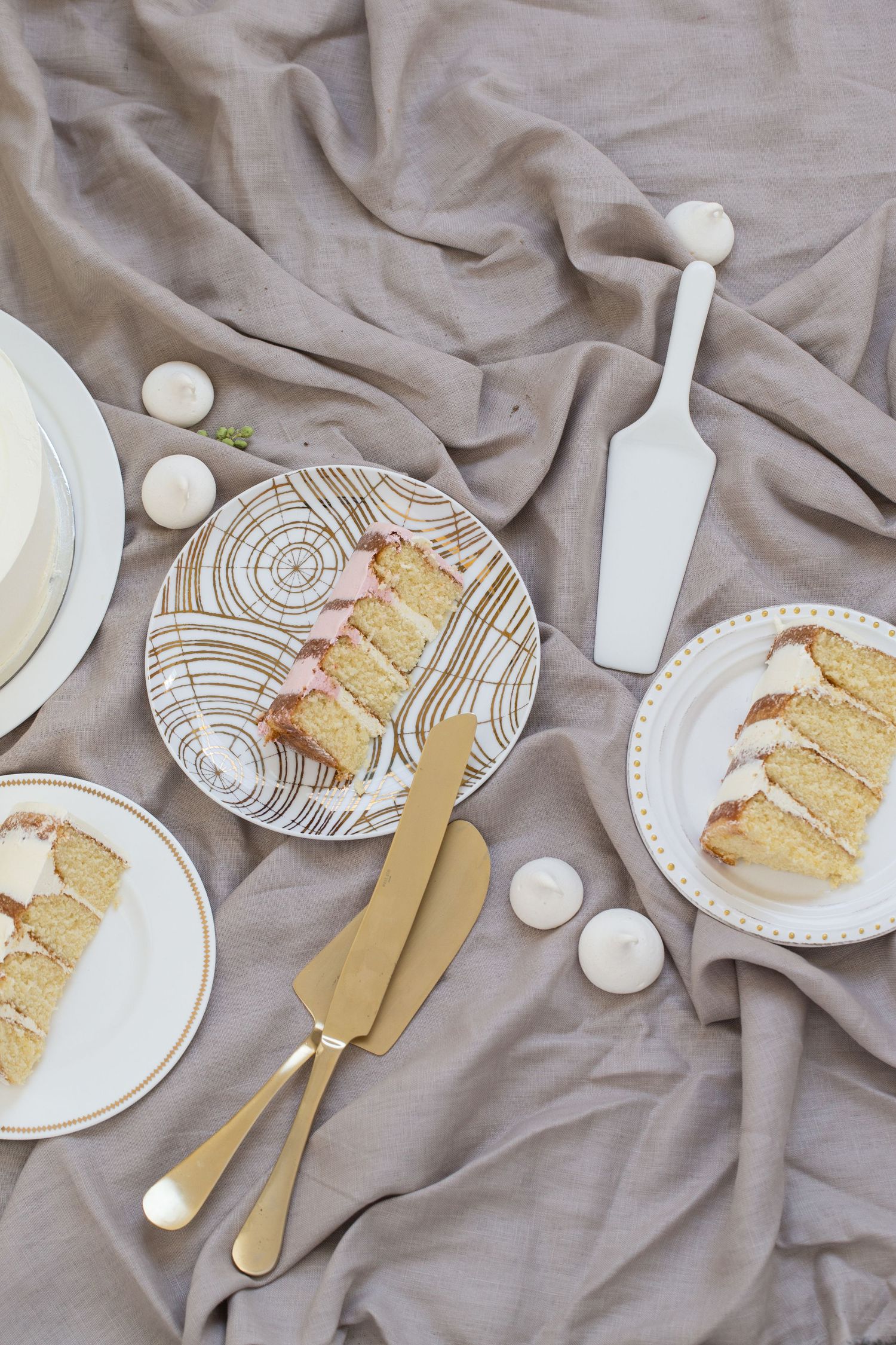 Elegant wedding cake slices on decorative plates with gold cutlery on grey linen fabric.