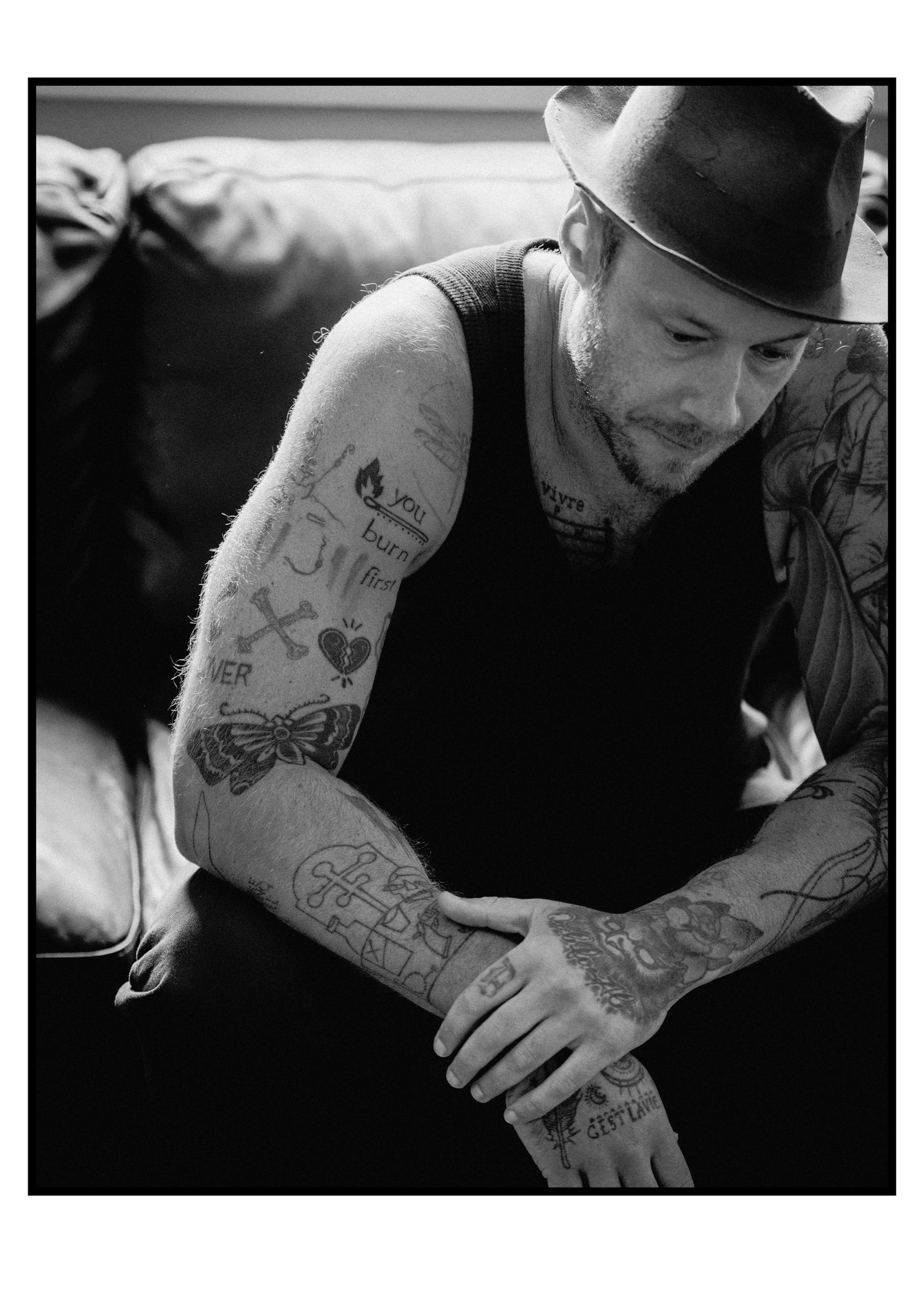 Tattooed man in black tank top and fedora hat sits on couch looking down at his ink-covered arms.