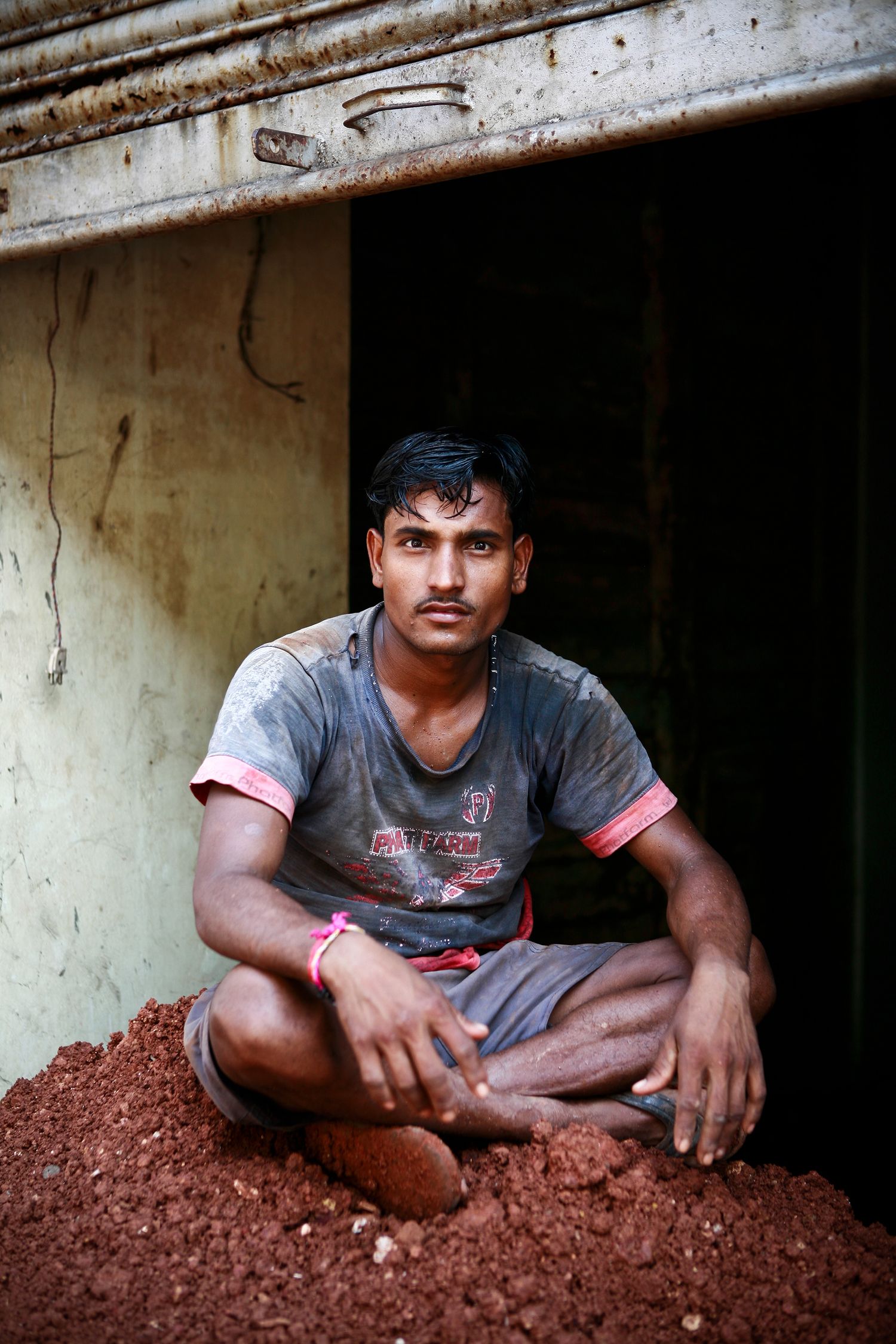 Young Indian male worker in dirty gray t-shirt sitting cross-legged on red soil in a doorway.