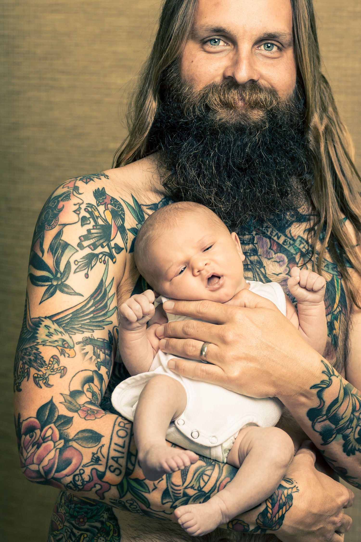 Heavily tattooed bearded man with long hair gently holding sleeping newborn baby.
