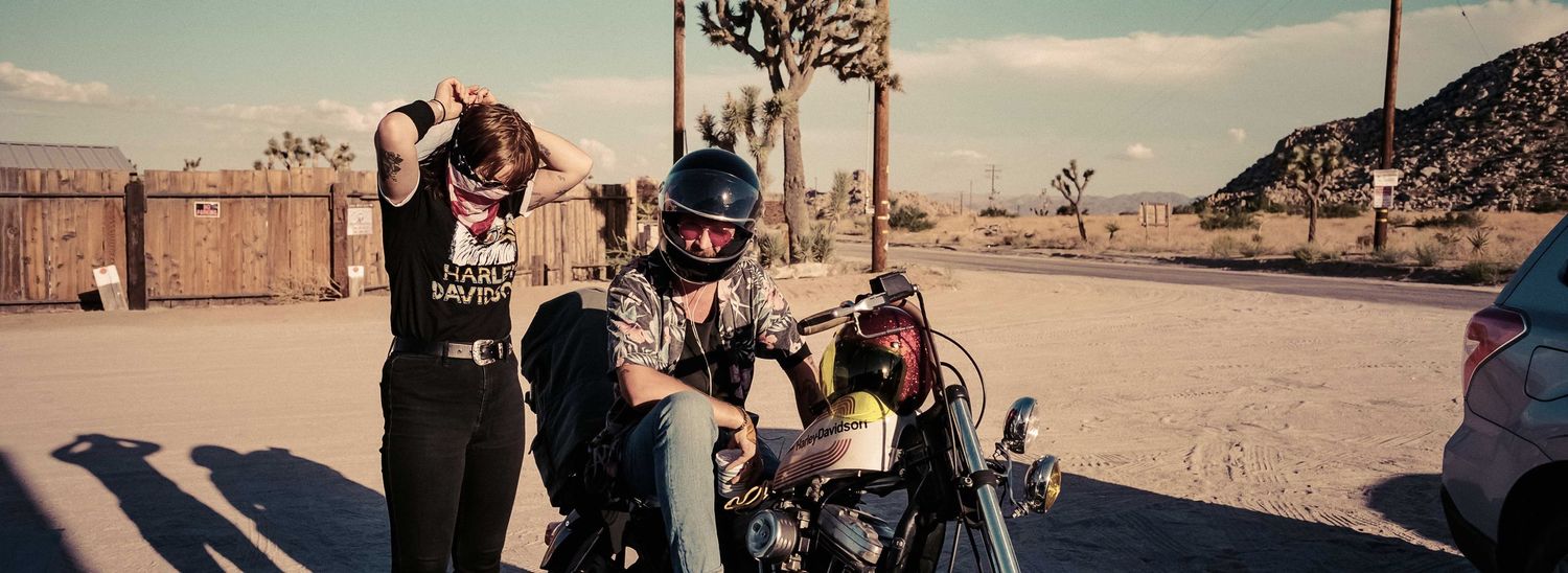 Two people with a vintage motorcycle in a desert landscape with Joshua trees.