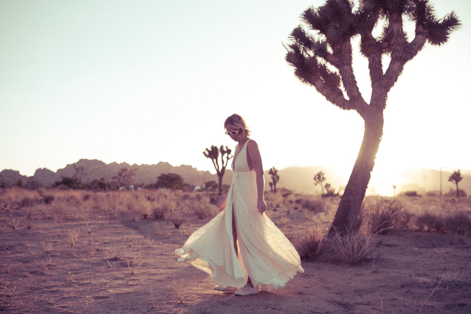 Bride in flowing white gown twirling near a Joshua Tree in a warm desert sunset landscape.