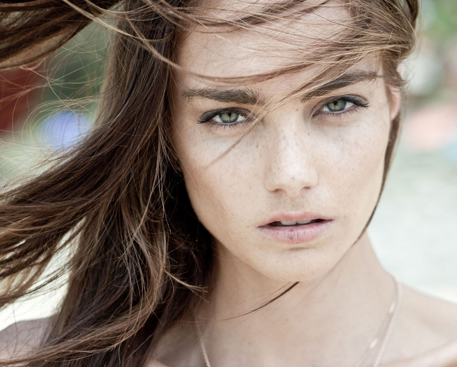 Close-up portrait of a young woman with green eyes, freckles, and windswept brown hair outdoors.