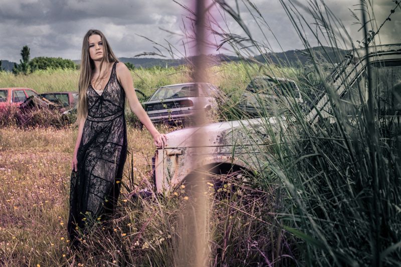 A woman in a long dark dress poses near abandoned vintage cars in an overgrown field.