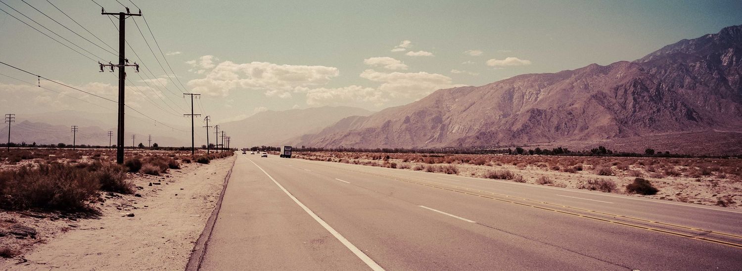 Empty desert highway stretching toward mountain range with telephone poles under hazy sky.
