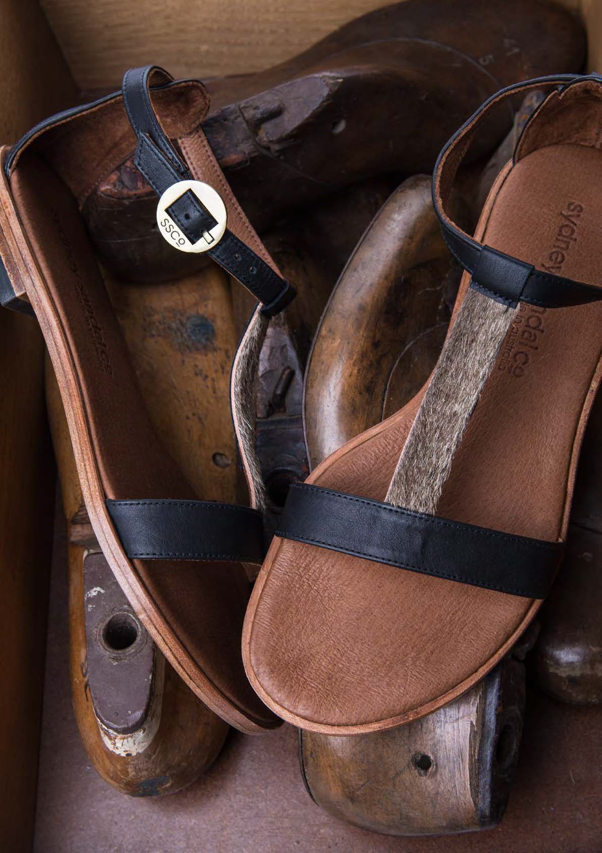 Black leather T-strap sandals with tan footbed displayed on rustic wooden shoe lasts.
