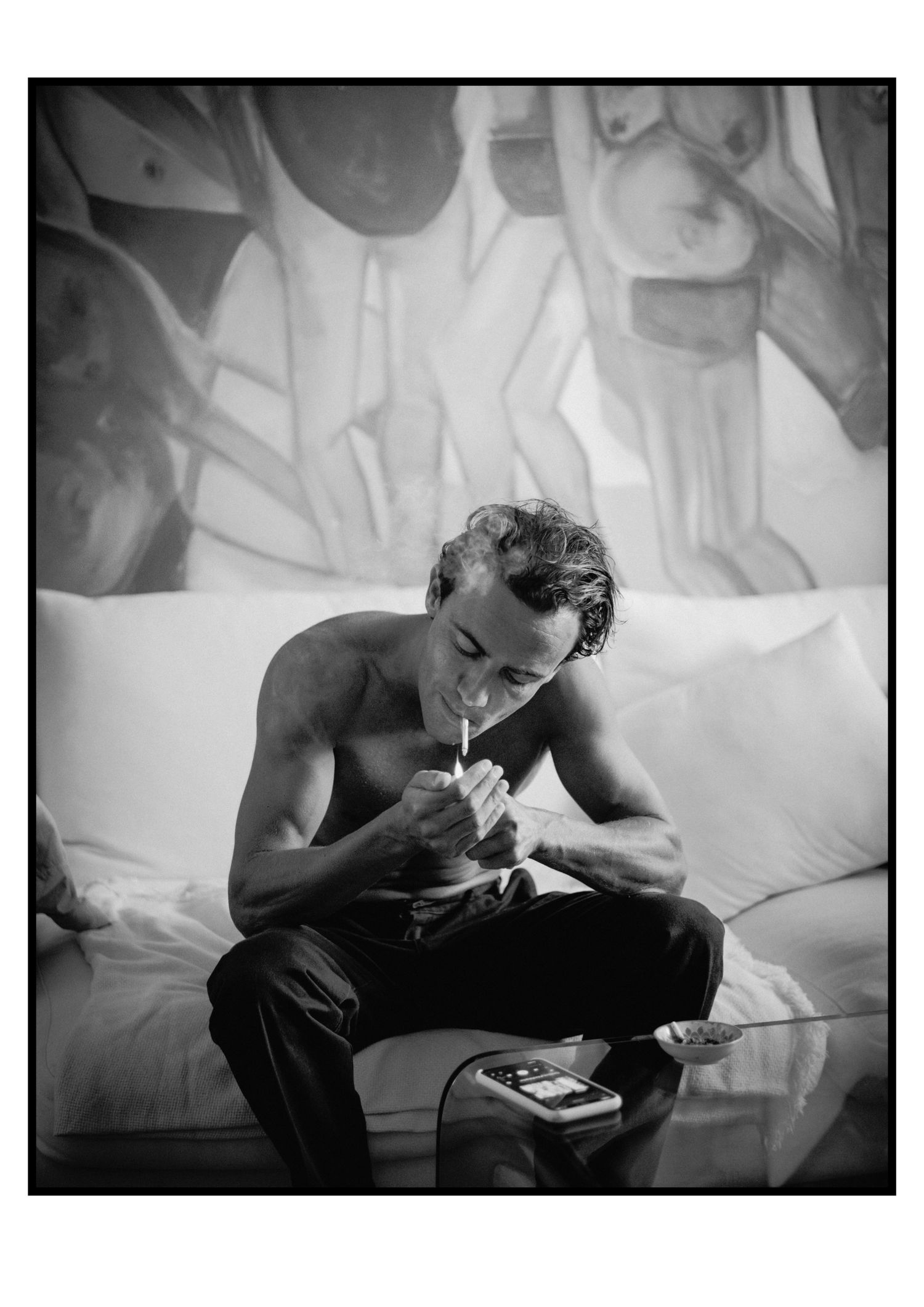 Shirtless man lights cigarette while seated on white couch beneath large figurative painting indoors.