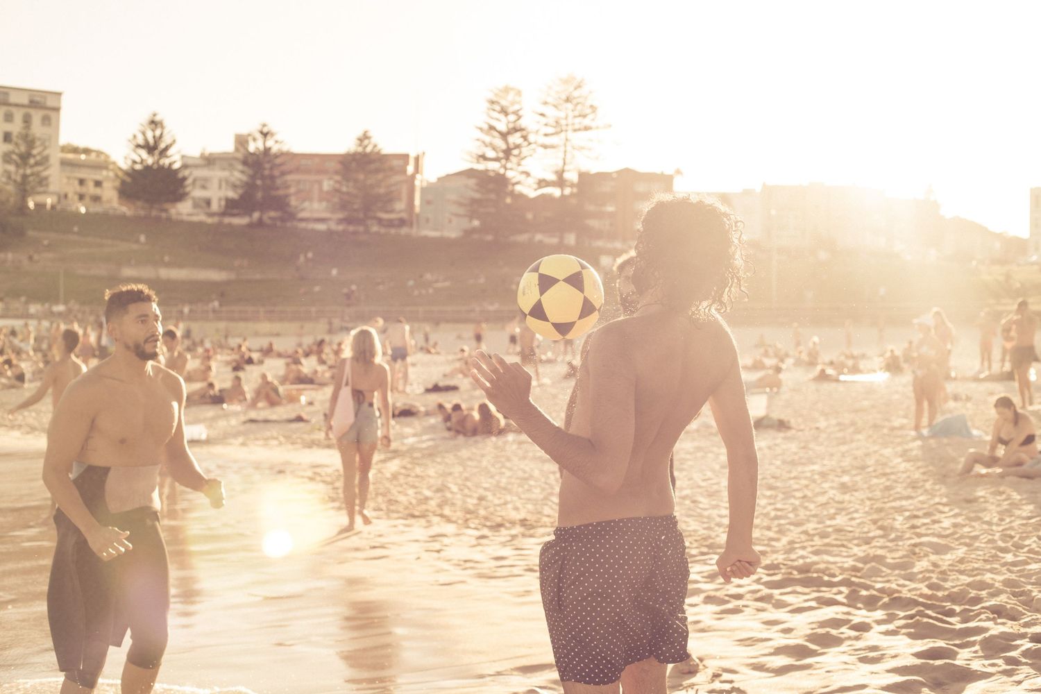 Two shirtless men playing with a soccer ball on a sunny crowded beach at golden hour.