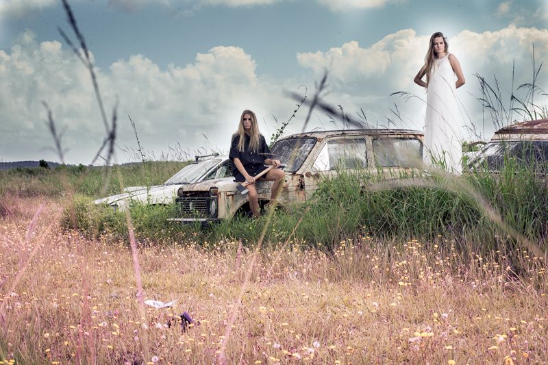 Two women pose with an abandoned rusty car in a wild flower field under dramatic clouds.