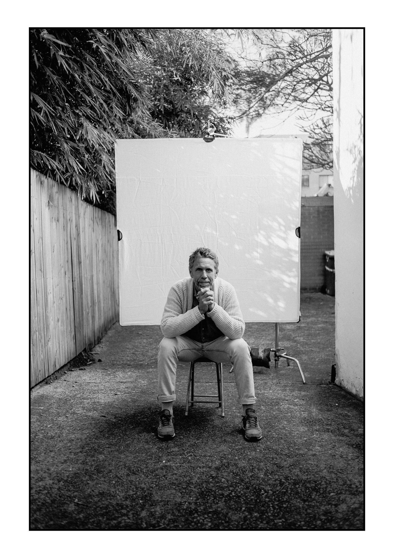 Man in light sweater sits on stool before white backdrop in outdoor alleyway, hands clasped thoughtfully.