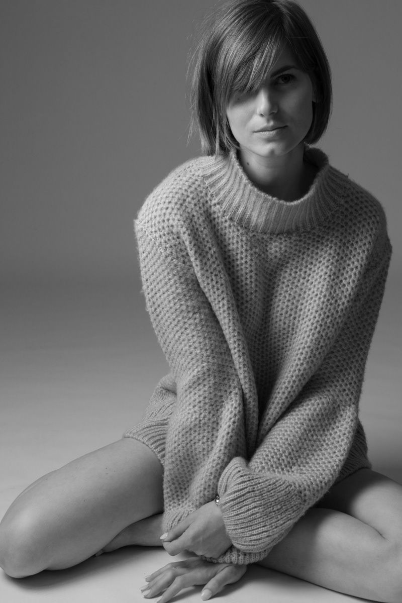 Black and white portrait of a woman with short hair sitting in an oversized knit turtleneck sweater.