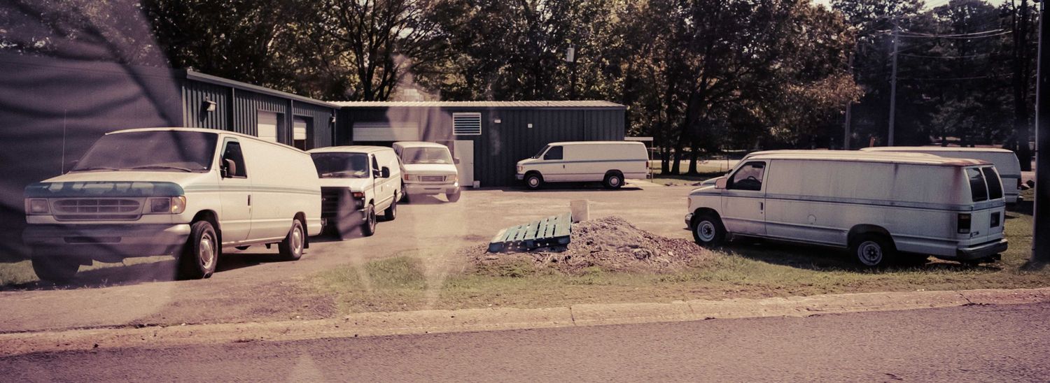 Vintage film photo of multiple white vans parked outside a suburban building on a sunny day.