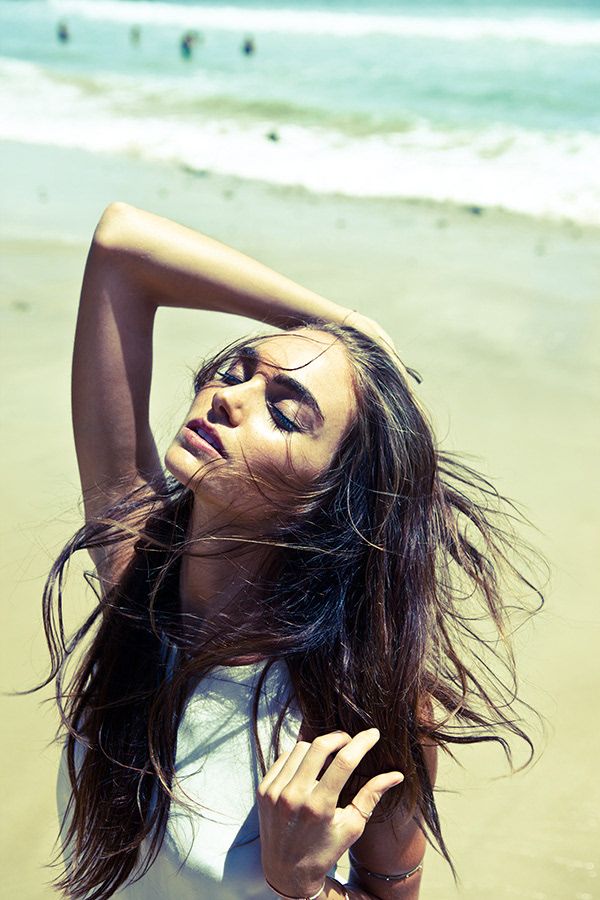 Young woman with long dark hair posing on a sunny beach with ocean waves in the background.