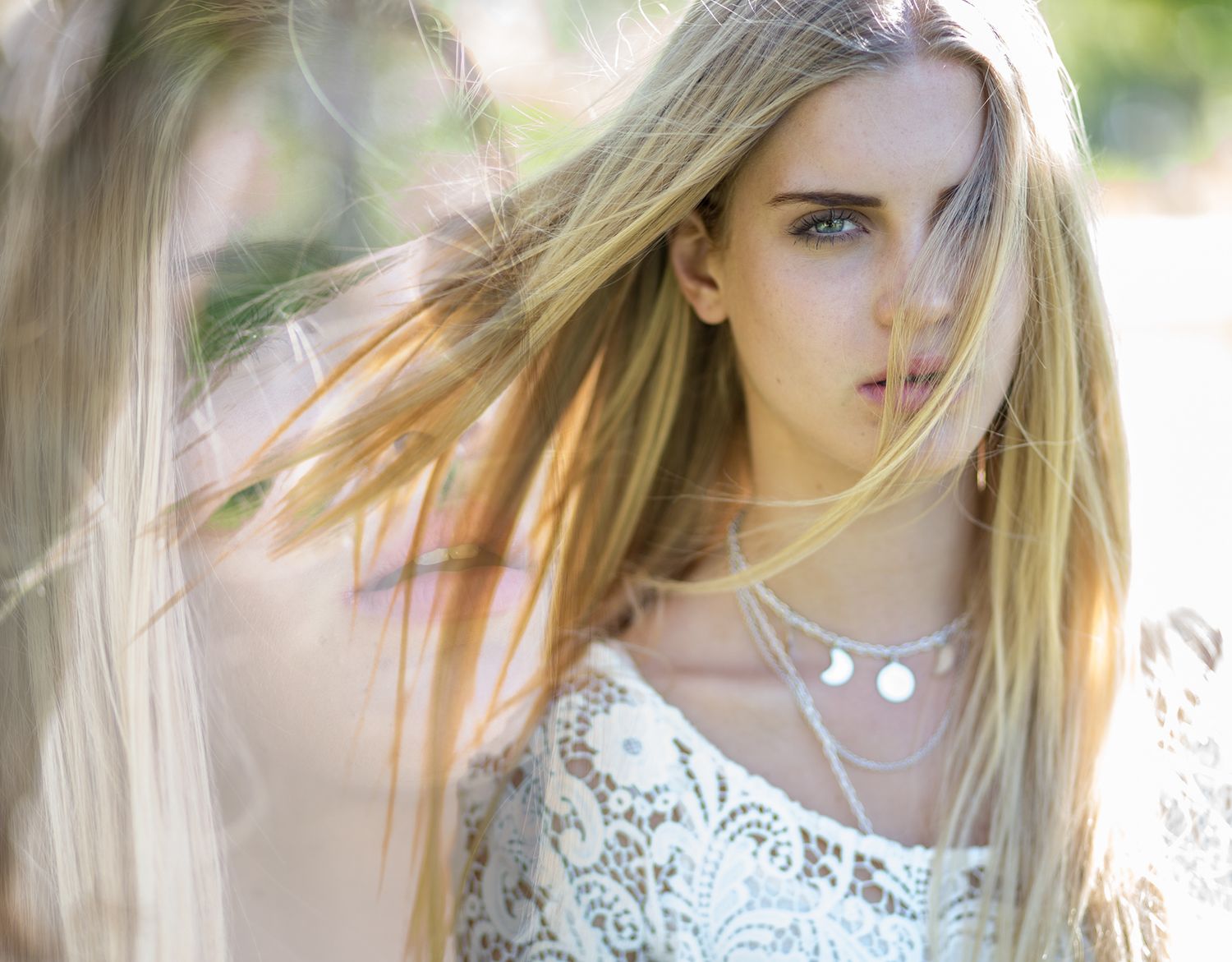 Close-up of blonde woman with blue eyes wearing lace top and layered necklaces in sunlit setting.