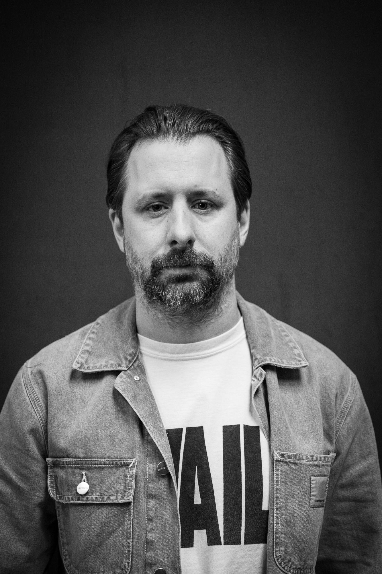 Black and white portrait of a bearded man in a denim jacket and graphic tee.