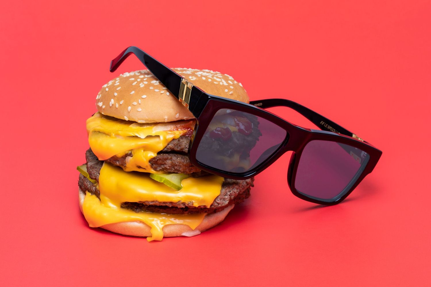 Stack of double cheeseburgers wearing black sunglasses on a bold red background.