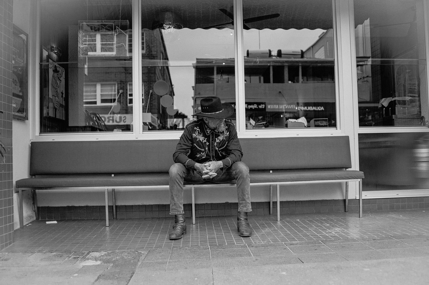 Man in cowboy hat sitting alone on bench outside storefront, black and white street photography.