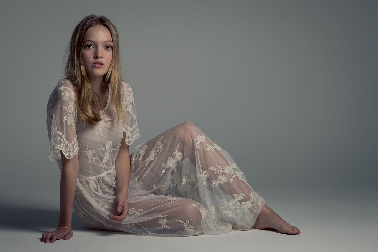 Young woman in a sheer white lace dress sitting gracefully on the floor against a neutral gray background.