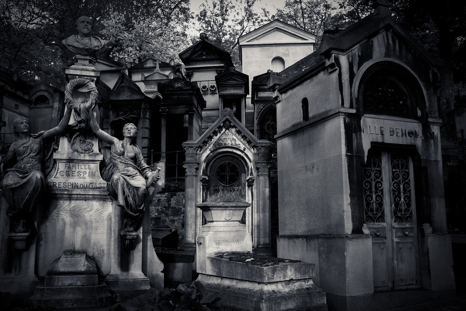 Dark gothic black and white photo of ornate cemetery mausoleums and stone sculptures.