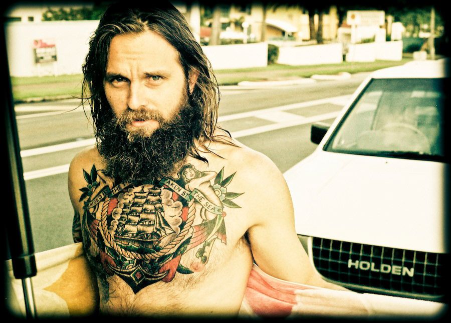 Portrait of a shirtless bearded man with chest tattoos leaning against a Holden vehicle.