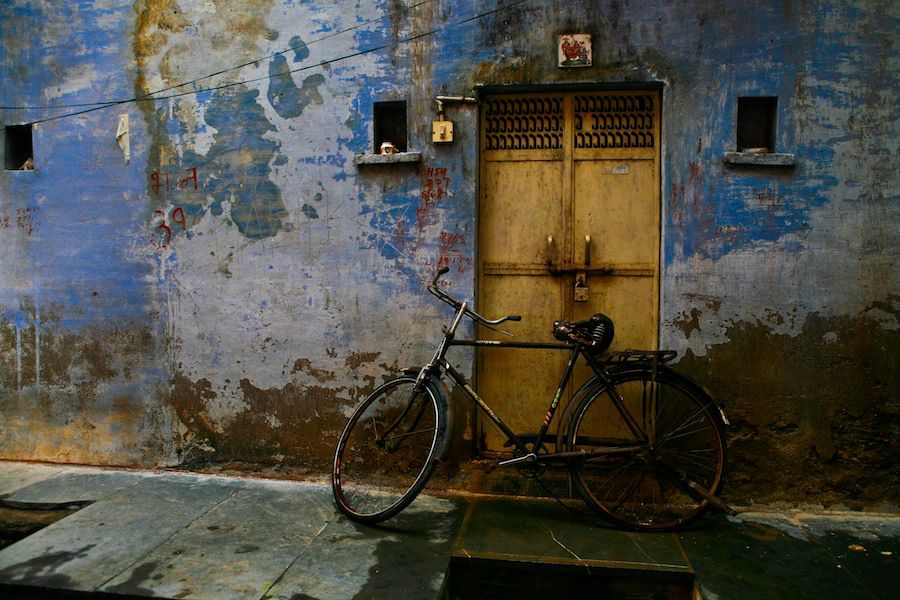 A vintage bicycle leans against a weathered blue wall beside an ornate wooden door in India.