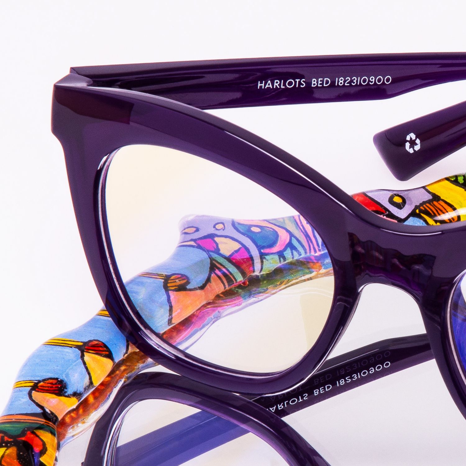 Close-up of purple cat-eye glasses frames labeled Harlots BED with colorful artistic temple design.
