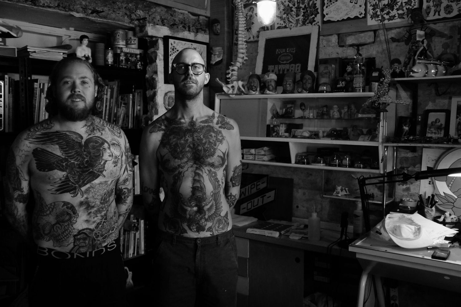 Two heavily tattooed men standing shirtless in a tattoo shop interior.