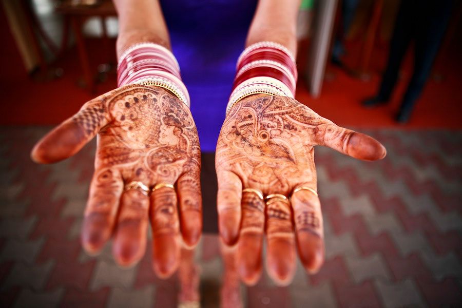 Close-up of hands adorned with intricate henna mehndi designs and red bangles at an Indian wedding ceremony.