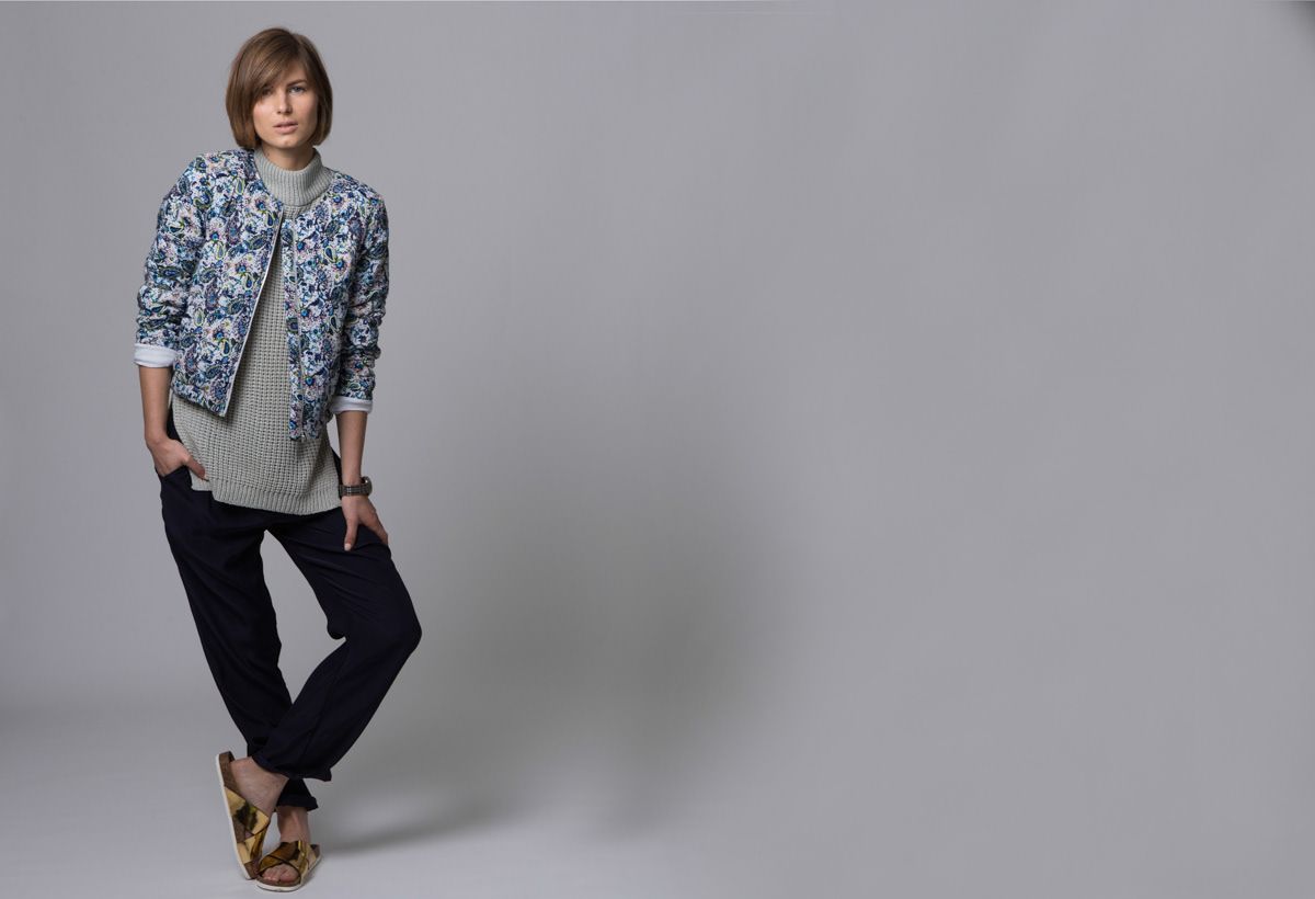A woman models a floral blue jacket with gray top and dark trousers on a gray background.