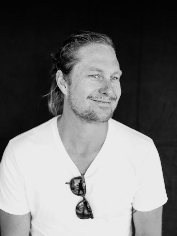 Smiling blond man with sunglasses tucked in shirt collar posing against dark background.