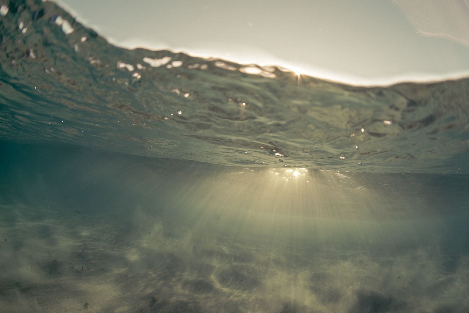 Serene underwater ocean view with sunlight rays filtering through a shallow wave above sandy floor.