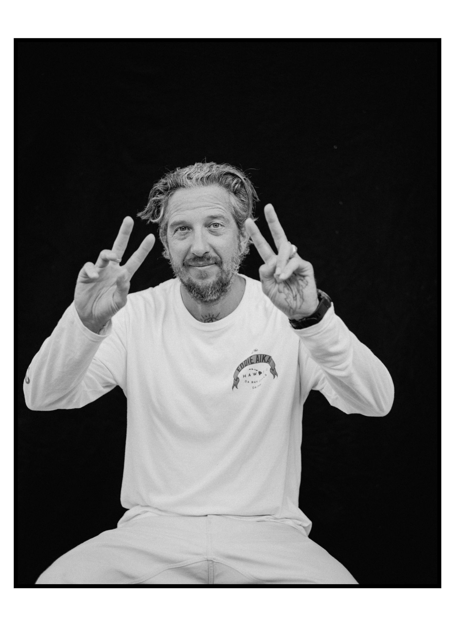 Bearded man in white sweatshirt makes peace signs with both hands in a black and white portrait photo.