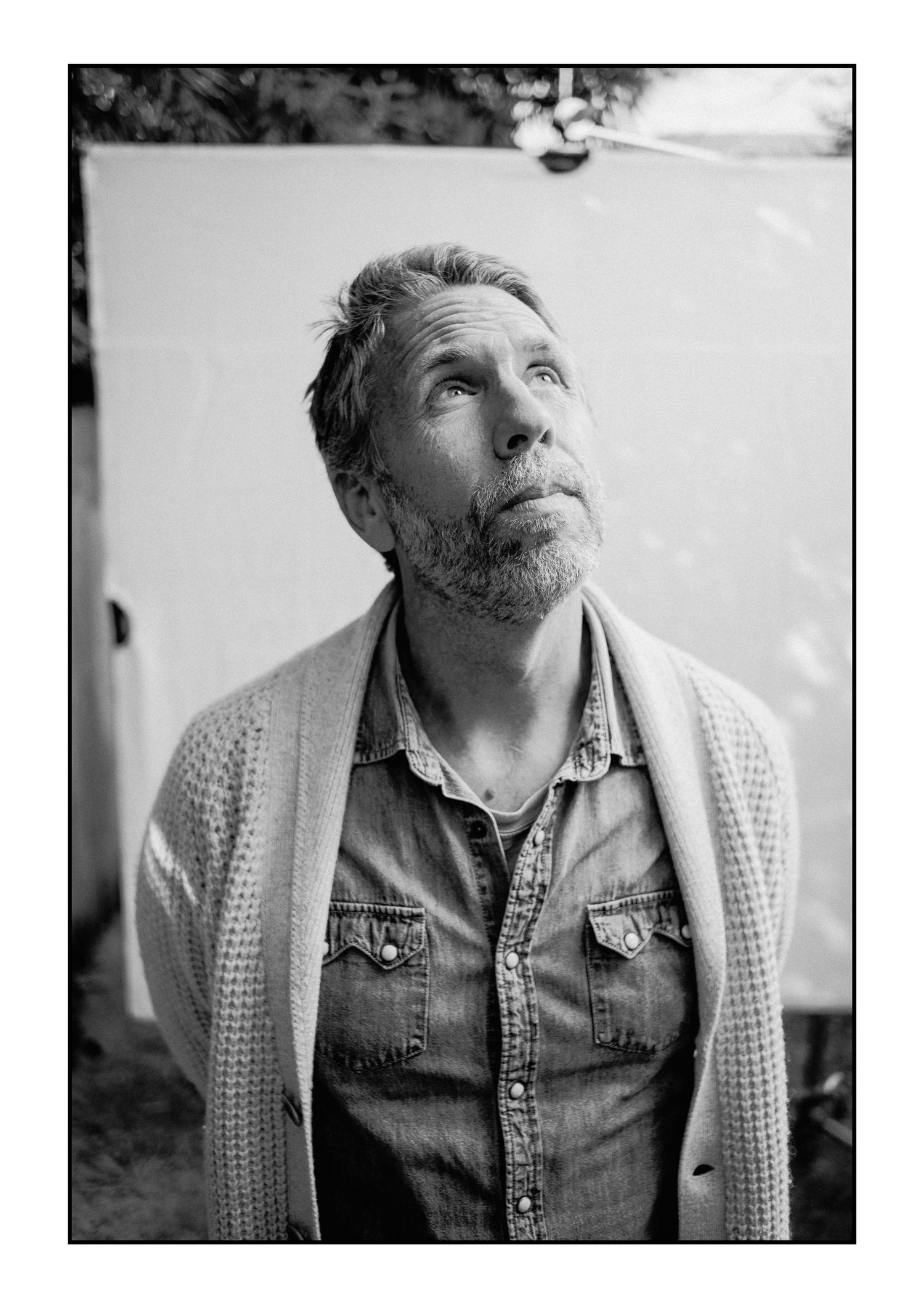 Middle-aged bearded man in denim shirt and cardigan looks upward outdoors in black and white portrait.
