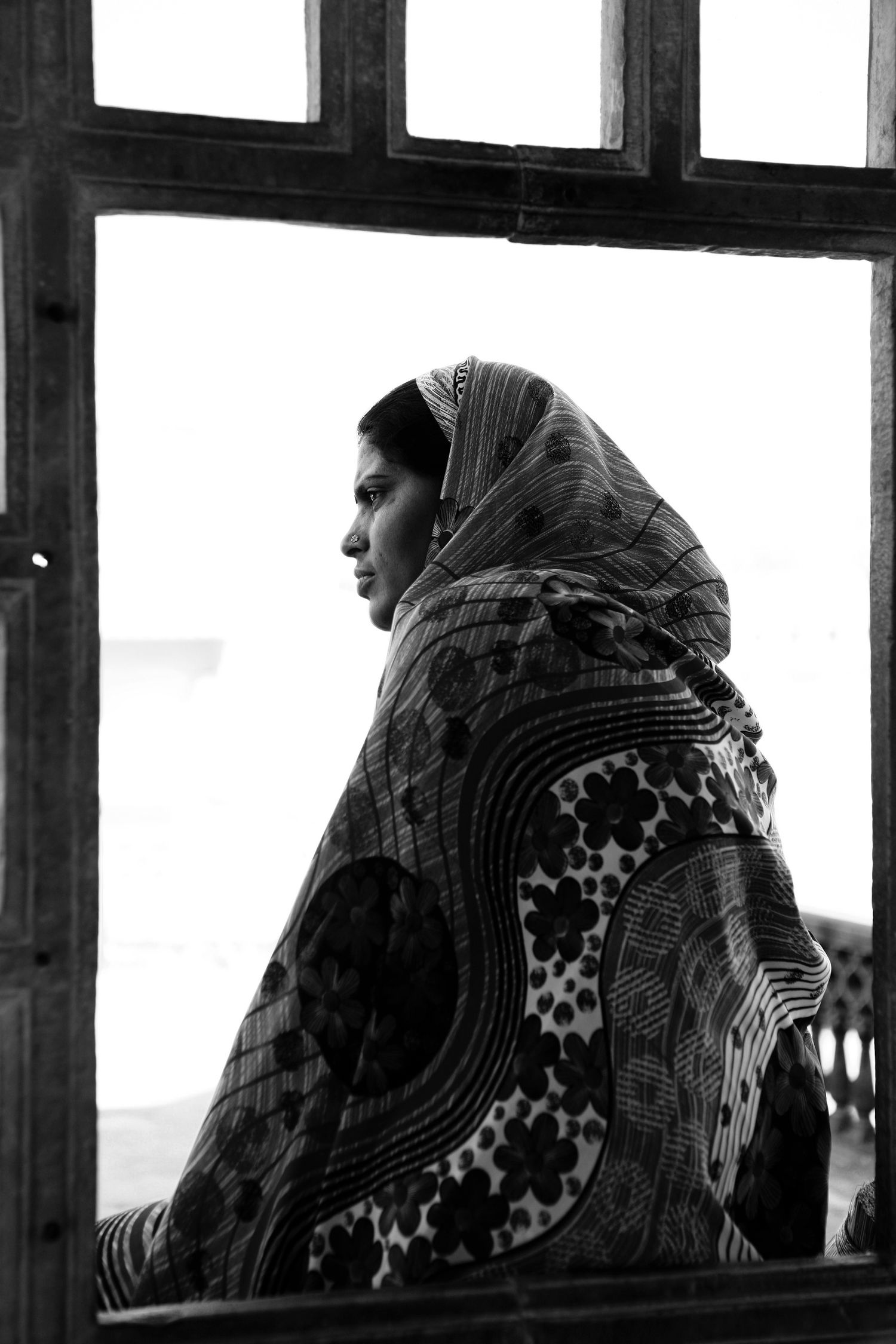 Black and white portrait of an Indian woman wrapped in a patterned shawl gazing through a wooden window frame.