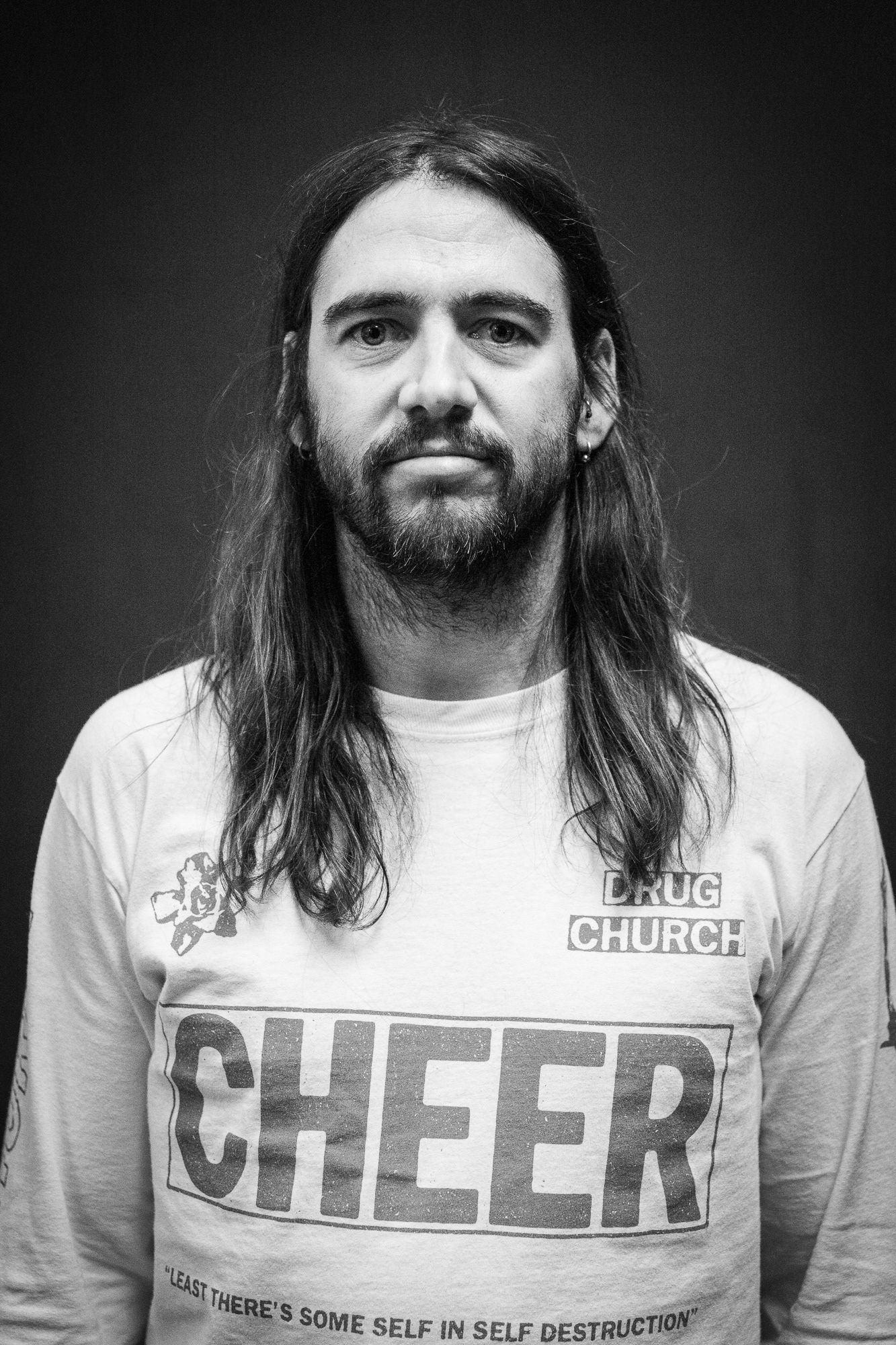 Black and white portrait of a long-haired bearded man wearing a Drug Church Cheer band t-shirt.