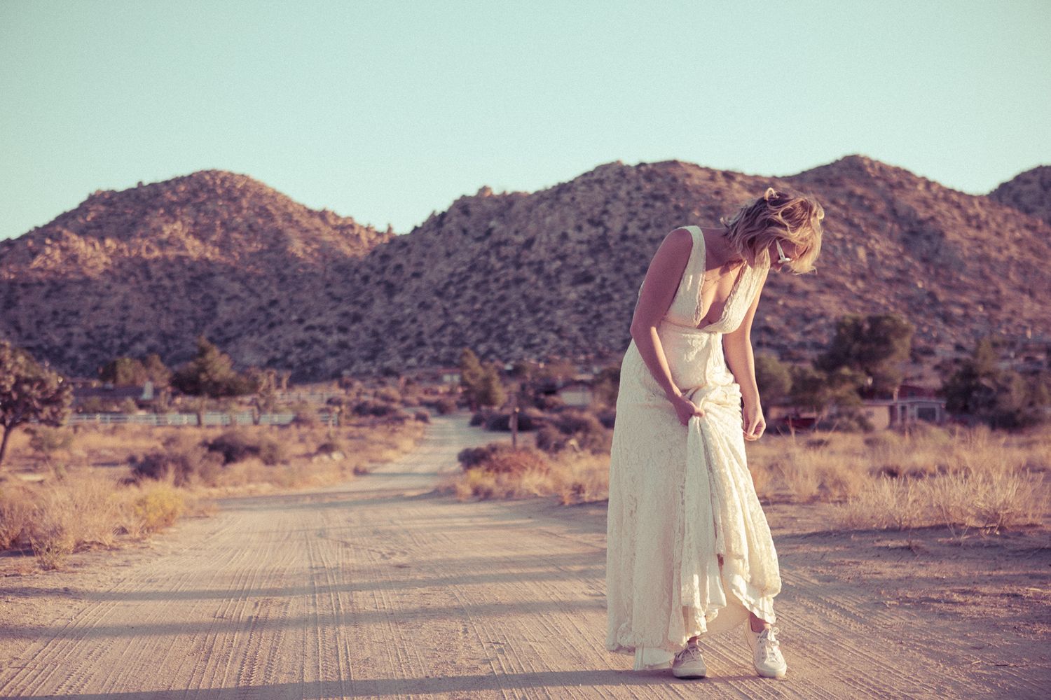 Woman in white dress standing on desert road with mountains in background.