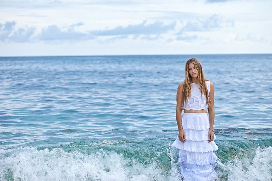 Young woman in white ruffled skirt set standing in ocean waves on beach.