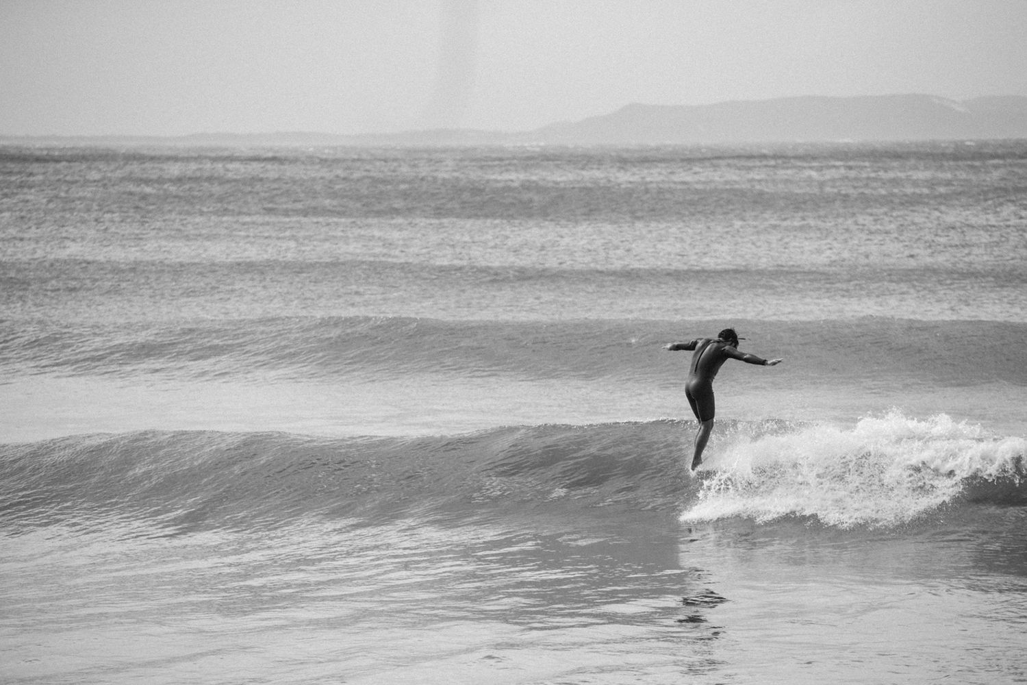 Black and white photo of surfer riding small ocean wave on beach.