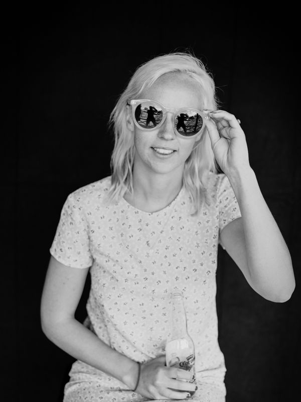 Black and white portrait of a smiling blonde woman holding sunglasses and a drink.
