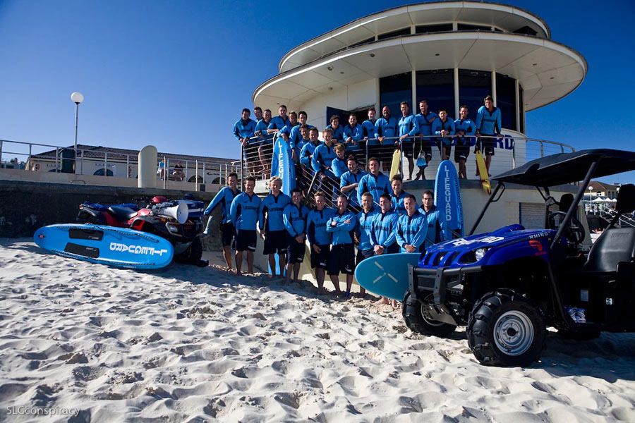 Group of lifeguards in blue uniforms posing on beach with rescue equipment.