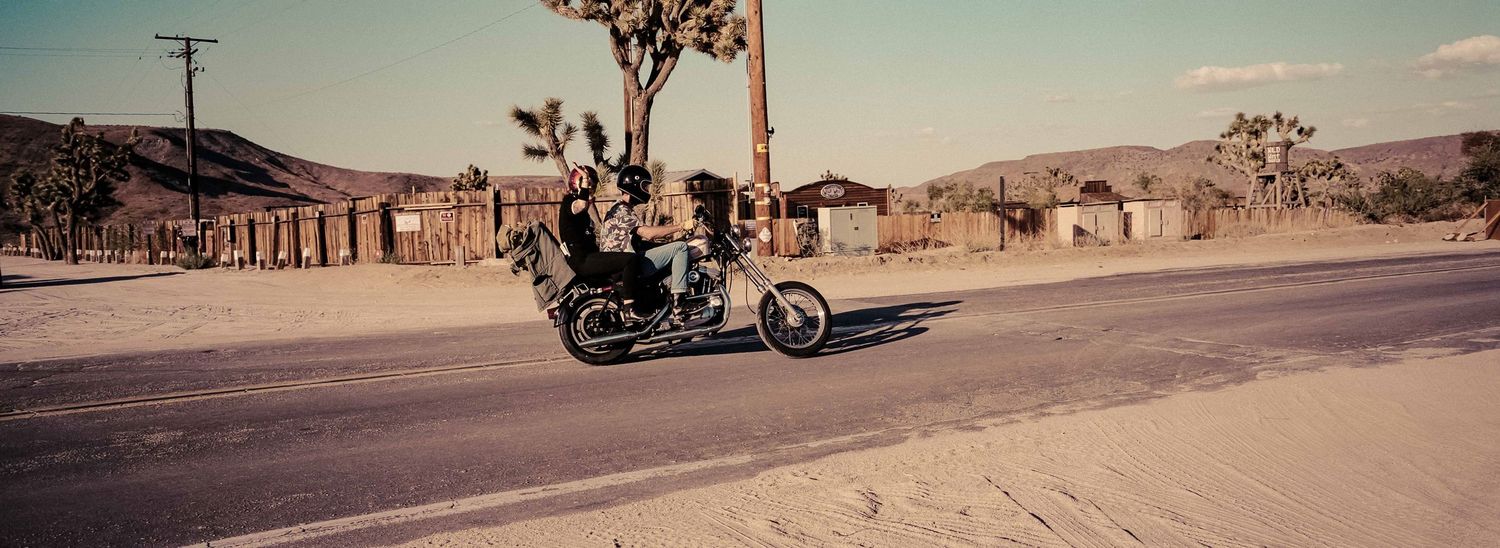 Motorcyclist riding along a desert highway past rustic buildings and Joshua trees in warm sunlight.