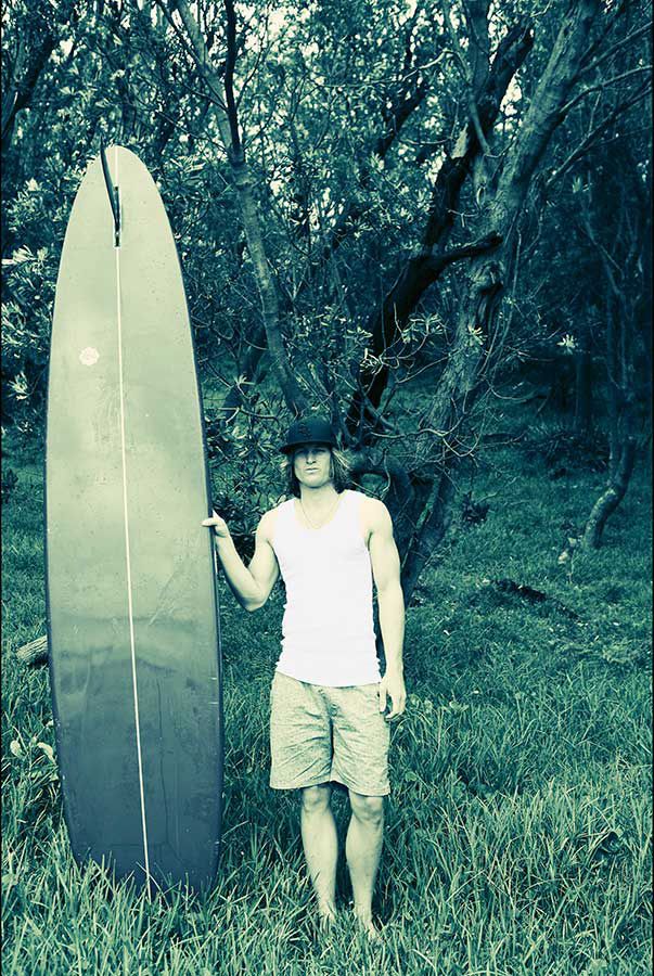 Person holding a longboard surfboard standing in a lush green forest clearing.